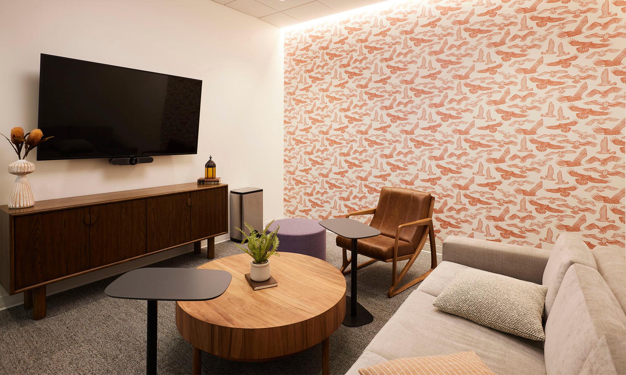 An office space with a couch, chair, table, and tv, behind an orange bird inspired wallpaper.