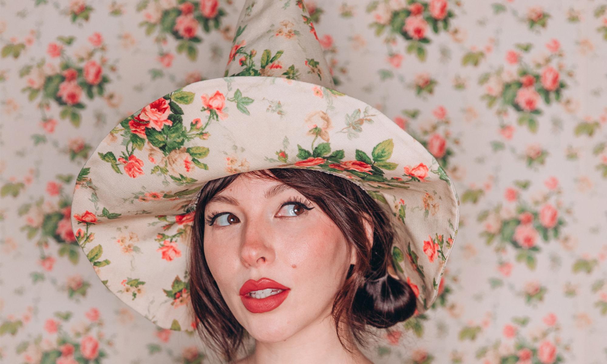 Keiko Lynn wearing a pink floral witch hat and dress in front of matching wallpaper.